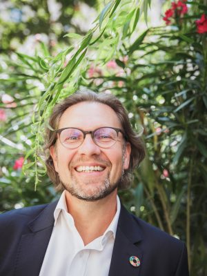 Michael Bauer-Leeb, Portrait vor einer großen grünen Pflanze, längere braune Haare, breites Lächeln, Brille