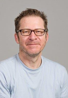 Jürgen Schneider mit schwarzer Brille. Graues Shirt, brauner Hintergrund. lächelnd