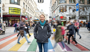 Gudrun Kirchert vor Regenbogenzebrastreifen, verschwommene gehende Menschen im Hintergrund, schwarze Jacke, blauer Schal